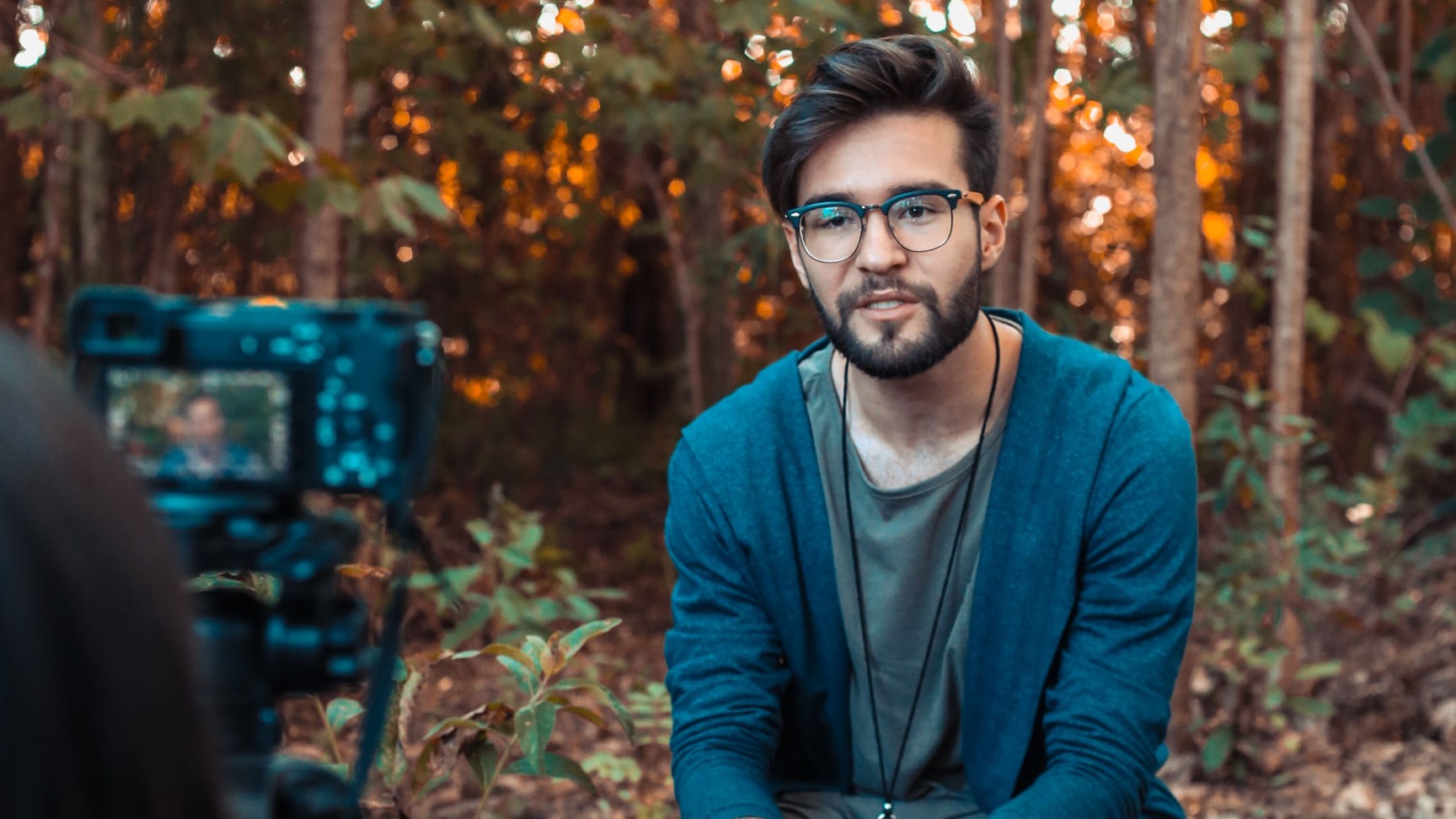 A man with dark hair, beard and glasses seated on a wooden stool in a sunlit wood, being filmed by a camera on a tripod in the foreground