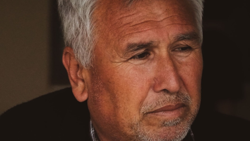 An older man with grey hair and stubble, deep in thought, in warm low light