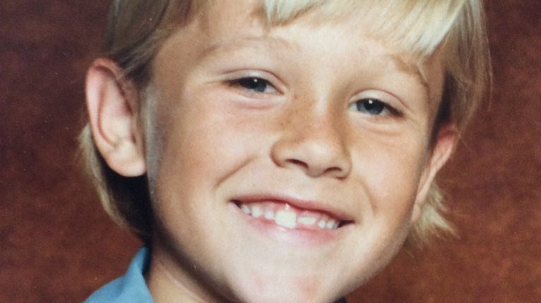 A smiling young boy with blond hair and freckles in a vintage studio portrait, against a mottled brown backdrop